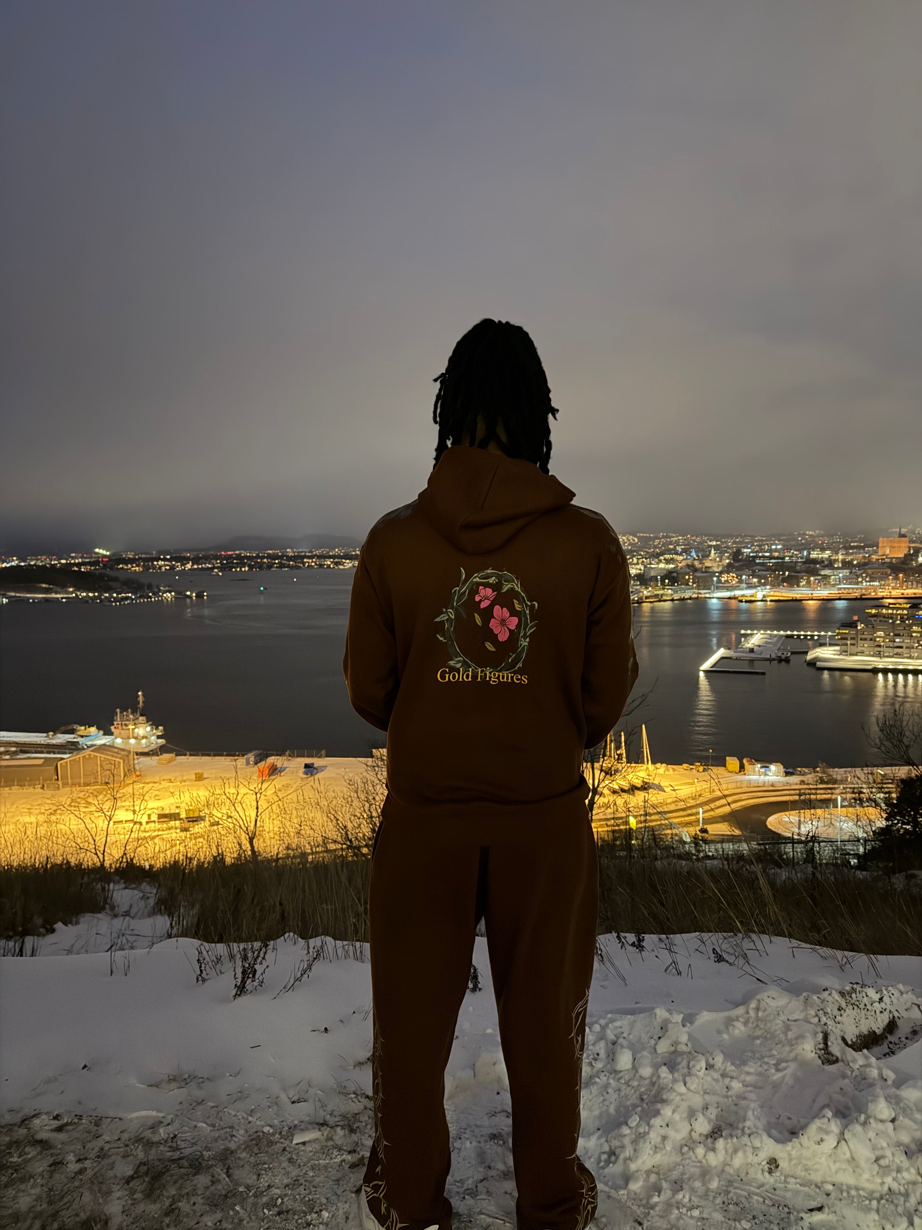 Person wearing a brown tracksuit with a floral design in a snowy landscape with illuminated buildings in the background.